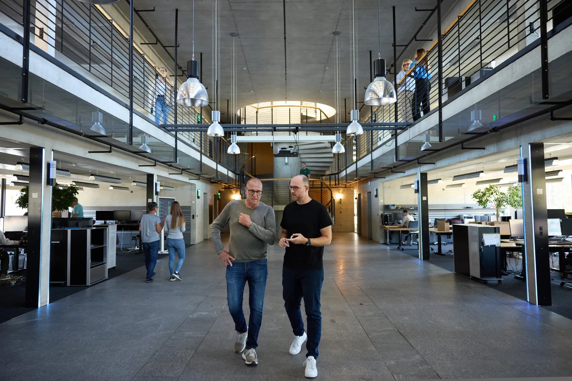 Zwei Mitarbeitende gehen durch die offene Bürohalle am Standort Seuzach, darüber verläuft die Galerie des Obergeschosses.